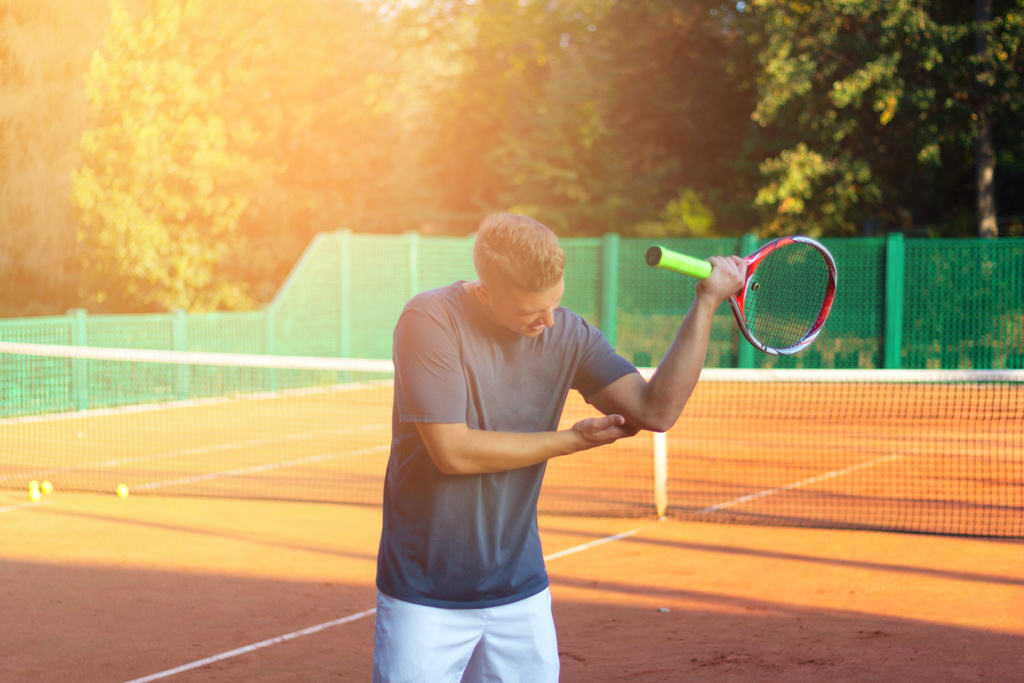 Handsome man on tennis court. Young tennis player. Pain in the elbow with sunlight yellow in background